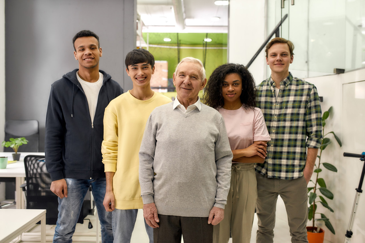 Happy diverse team of employees looking at camera while posing with senior intern, standing together in the office, Age diversity in the workplace