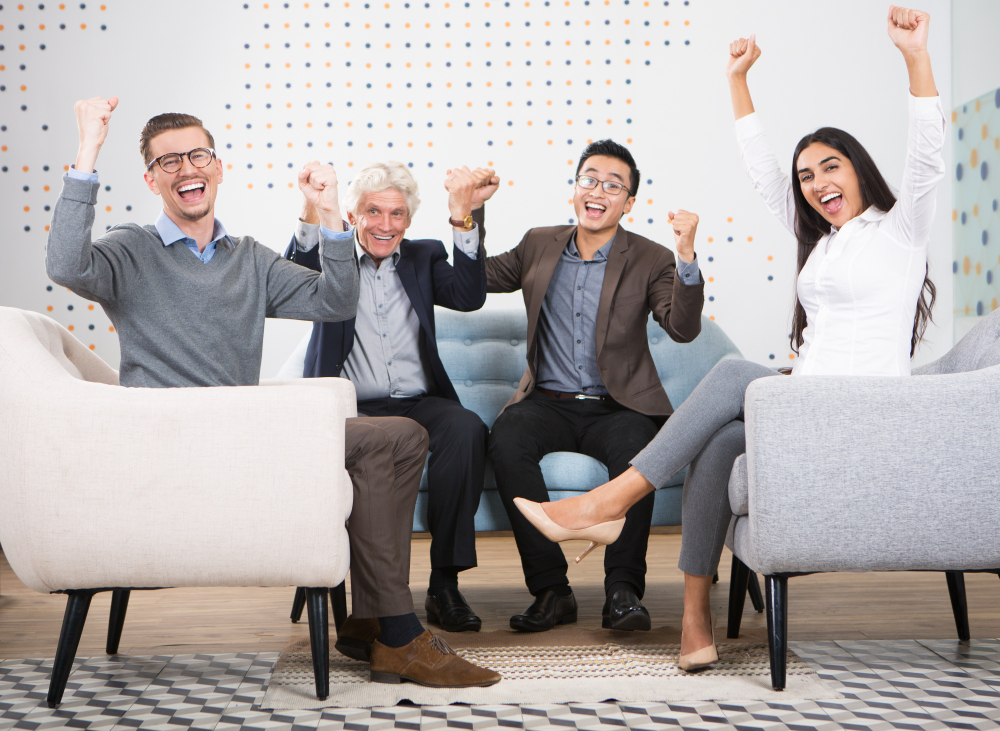 Diverse business people sitting on grey couch celebrating success with fists up in the air